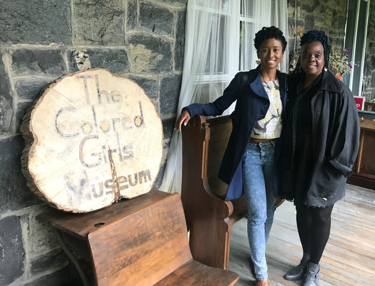 Two Black women stand next to each other to the right side of a wooden sign stating "The Colored Girls Museum"